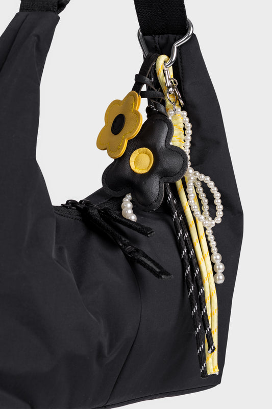 mustakeltainen kukkalaukkukoru kuvattuna mustassa laukussa. Lisänä korussa myös kaksi roikkuvaa nauhaa ja helmikoriste.